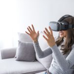 Young elegant woman sitting on sofa chair wearing virtual reality goggles looking online website and using hand moving 3D simulation screen. Enjoying new reality. Attractive young woman in VR headset