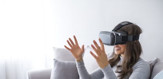 Young elegant woman sitting on sofa chair wearing virtual reality goggles looking online website and using hand moving 3D simulation screen. Enjoying new reality. Attractive young woman in VR headset
