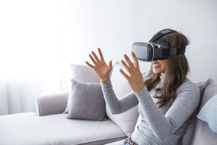 Young elegant woman sitting on sofa chair wearing virtual reality goggles looking online website and using hand moving 3D simulation screen. Enjoying new reality. Attractive young woman in VR headset